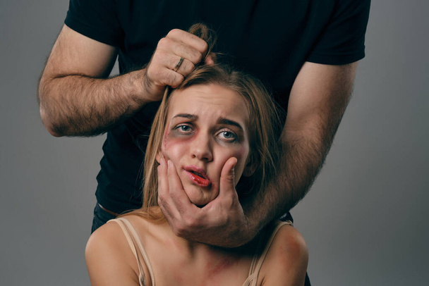Strong man is holding by hair scared female with tousled hair and bruises on her face. Gray studio background. Domestic violence, abuse. Close-up. - Photo, Image