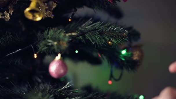 Close-up of the hand of a little child girl decorates a New Year tree. Hangs toys on branches against the background of garlands and lights - Filmagem, Vídeo