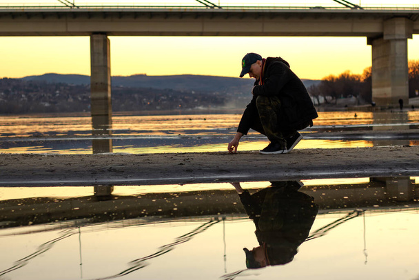  Troubled young man with baseball cap in squat, under the bridge on the sandy river bed after the river has dried up after a dry winter. Sunset. Reflection in the water. - Photo, Image