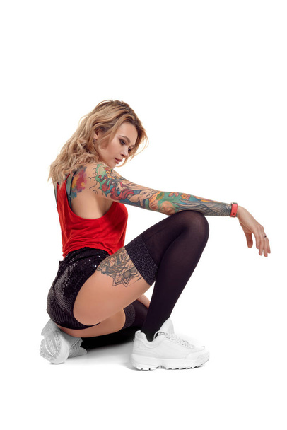 Σέξι ξανθιά γκόμενα με τατουάζ και μακριά σγουρά μαλλιά ποζάρει στο στούντιο. - Φωτογραφία, εικόνα