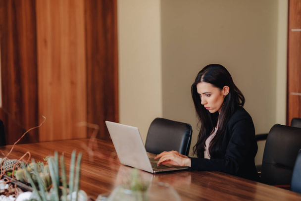 Menina emocional atraente jovem em roupas de estilo empresarial sentado em uma mesa em um laptop e telefone no escritório ou auditório sozinho
 - Foto, Imagem