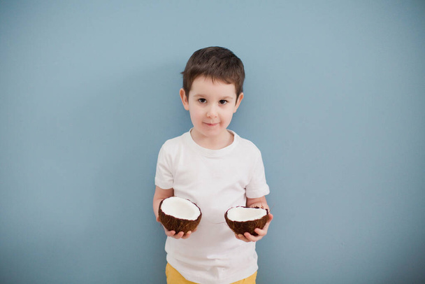 Little boy in white t-shirt and yellow shorts holding two coconuts on the background of the blue wall - Photo, Image