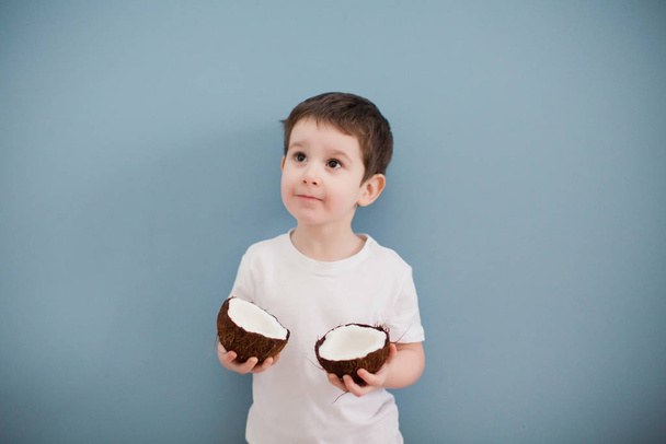 Kleine jongen in wit t-shirt en gele broek met twee kokosnoten op de achtergrond van de blauwe muur - Foto, afbeelding