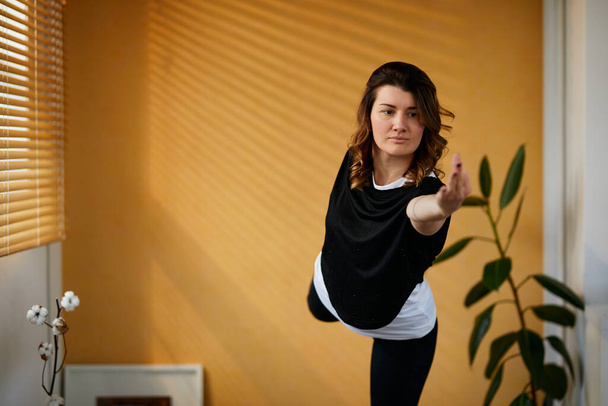 Полная длина тонкой кавказской женщины-йога, стоящей на одной ноге в положении "Повелитель танца" и балансирующей. Интерьер дома, утреннее время
. - Фото, изображение