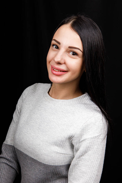 Portrait of serious beautiful female with black hair, has minimal makeup, looks calmly at camera, wears white jumper, stands against black background, being deep in thoughts - Photo, Image
