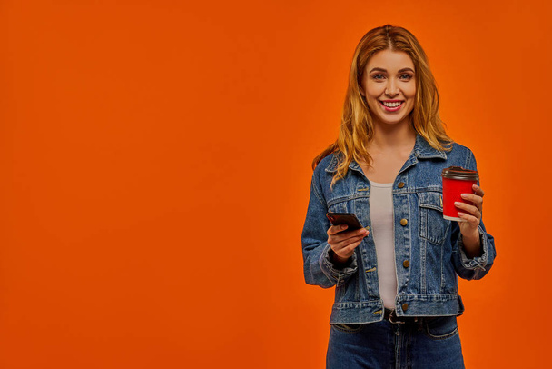 Glad woman holds the phone and a red cardboard cup of drink in her hands. - Photo, Image