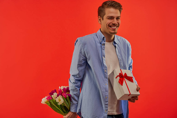 A guy with wide smile with white box with a red bow and a bouquet of flowers in hands. - Photo, Image