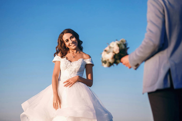 Nevěsta a ženich stojí u řeky proti modré obloze, muž nese kytici bílých růží a dává své ženě. usmívající se bruneta, designové svatební šaty, krásná svatba - Fotografie, Obrázek