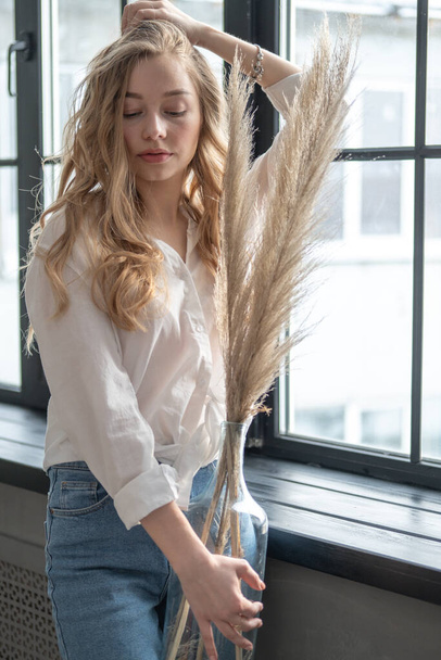 Young beutiful blond woman wearing white shirt and blue jeans standing next to window. Morning portrait of a girl holding vase with dry plants. - Photo, image