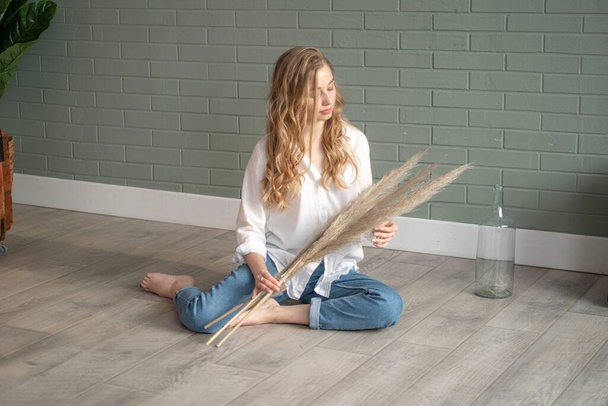 Young beautiful blond woman wearing white shirt and blue jeans sitting on the floor with bare feet next to vase with dry plants. Girl sitting in a daylight from the window. Copy space - Фото, изображение