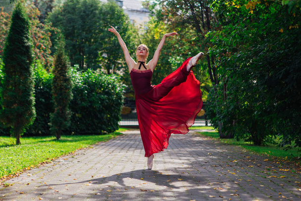 Woman ballerina in red ballet dress dancing in pointe shoes in autumn park. Ballerina standing in beautiful ballet pose - Foto, afbeelding