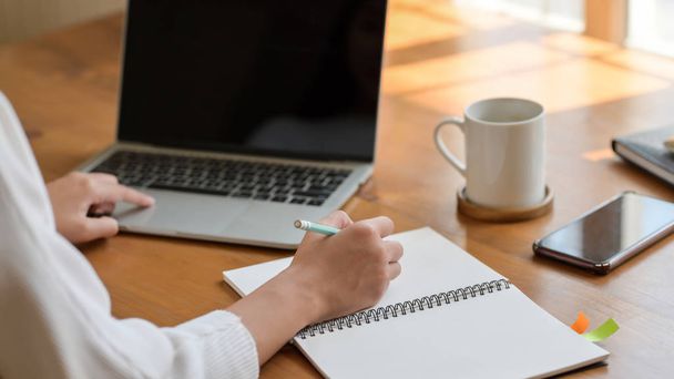 Close-up bekijken van jonge college meisje werken op laptop tijdens het schrijven op notebook met koffiebeker en smartphone in eenvoudige werkruimte  - Foto, afbeelding