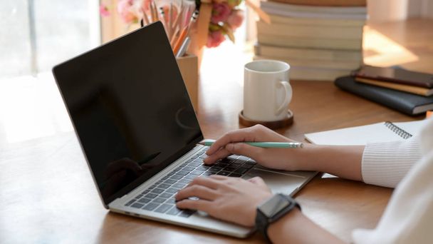 Обрезанный снимок молодой студентки, печатающей на ноутбуке во время работы над проектом с канцелярскими принадлежностями, книгами и кофейной чашкой на деревянном столе
  - Фото, изображение