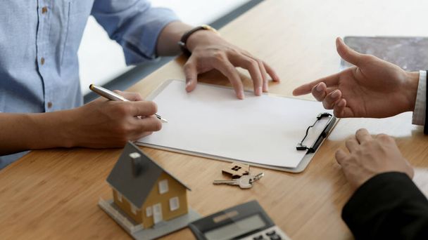 Περικοπή πλάνο του πελάτη ακινήτων υπογραφή συμφωνία δανείου σπίτι με το μοντέλο του σπιτιού και το κλειδί του σπιτιού στο ξύλινο γραφείο - Φωτογραφία, εικόνα