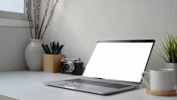 Colpo ritagliato del posto di lavoro moderno con computer portatile a schermo bianco, fotocamera e decorazioni sulla scrivania bianca accanto alla finestra
  - Foto, immagini