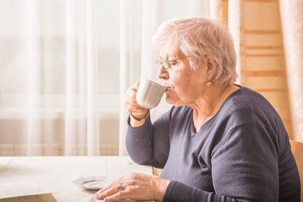 Yaşlı kadın evde sıcak bir içecek içme. Üst düzey emekli kadın holding fincan kahve ellerinde - Fotoğraf, Görsel