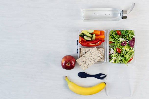 Vegetables and fresh green salad in lunch box, banana and apple on wooden white desk. Healthy business lunch at workplace top view. Copy space. - Photo, Image