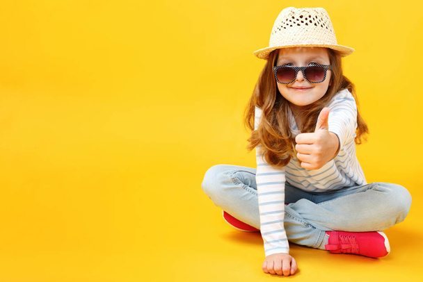 Little girl sitting on the floor in the studio with a hat and sunglasses on a yellow background. The child shows a thumb up. - Foto, Imagen