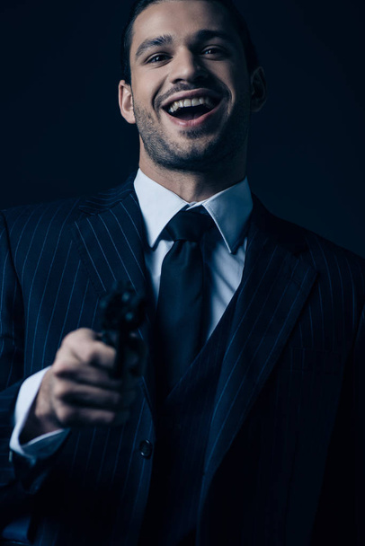 Selective focus of gangster aiming gun, laughing and looking at camera isolated on dark background - Photo, Image