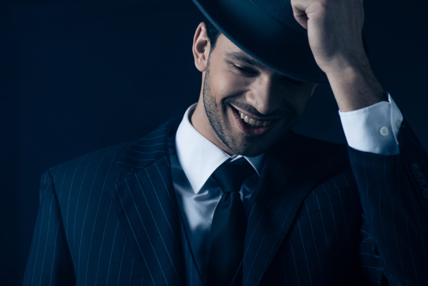 Front view of mafioso holding felt hat and smiling on dark blue - Foto, afbeelding
