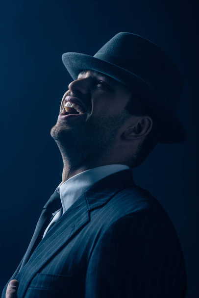 Mafioso in suit and felt hat laughing on dark blue background - Fotoğraf, Görsel