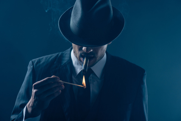 Mafioso with covered eyes with felt hat lighting cigar on dark blue background - Photo, Image