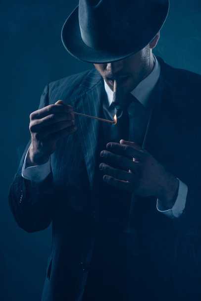 Mafioso with covered eyes with felt hat lighting cigar on dark blue  - Фото, изображение