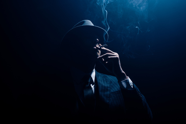 Silhouette of mafioso in suit and felt hat smoking cigarette on dark blue background - Фото, изображение