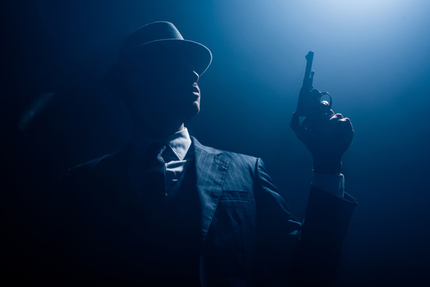 Vista de ángulo bajo de la silueta del gángster sosteniendo la pistola y fumando sobre fondo oscuro
 - Foto, imagen