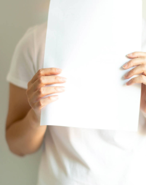 Close-up of a woman in a white T-shirt holding blank white paper; for fill a message or an advertise sentence. concept of communication and marketing.    - Photo, Image