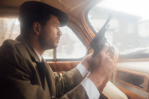 Profiel van mafioso in platte kap en jas met pistool rijden auto - Foto, afbeelding