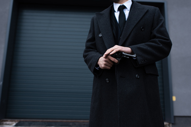 Cropped view of gangster in black coat holding revolver on street - 写真・画像
