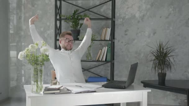 joyful man is holding hands behind head while sitting on chair near table with a computer in an office with a modern floral interior on background of fresh bouquets of flowers, happy male employee is - Video