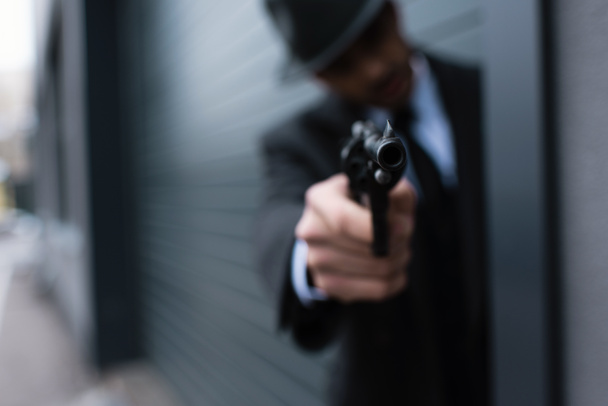 Selective focus of mafioso silhouette with outstretched hand aiming weapon from corner on street - 写真・画像