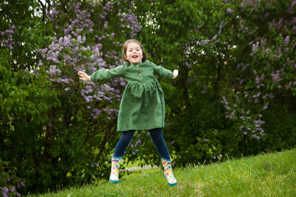 Roztomilá dívka v zeleném plátně šaty má zábavu v parku s kvetoucími šeříky, má jaro a teplo. Krásná jarní zahrada. Šťastné dětství, mír a štěstí koncept - Fotografie, Obrázek