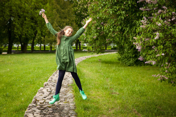 Nettes Mädchen in grünem Leinenkleid hat Spaß im Park mit blühenden Fliedern, genießt Frühling und Wärme. Schöner Frühlingsgarten. Glückliche Kindheit, Frieden und Glück - Foto, Bild