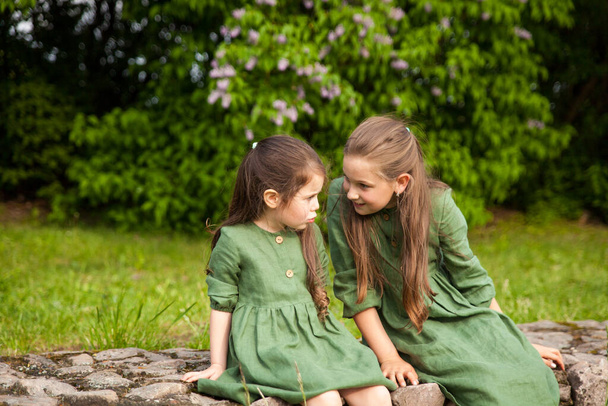 Дві сестри в зеленій лляній сукні розважаються в парку з квітучим бузком, насолоджуються весною і теплом. Прекрасний весняний сад. Щасливе дитинство. Концепція сім'ї, любові, миру та щастя
 - Фото, зображення