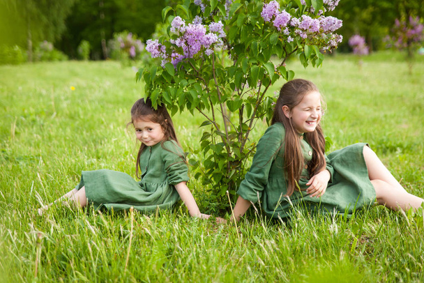 Zwei Schwestern im grünen Leinenkleid amüsieren sich im Park mit blühenden Fliedern, genießen Frühling und Wärme. Schöner Frühlingsgarten. Glückliche Kindheit. Familie, Liebe, Frieden und Glück - Foto, Bild
