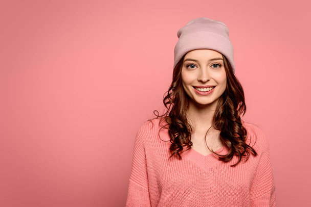 attractive cheerful girl in sweater and hat smiling at camera isolated on pink - Fotografie, Obrázek