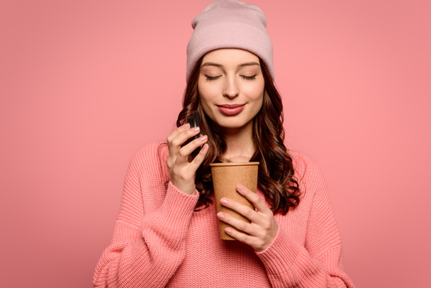 smiling girl enjoying flavor of coffee while opening disposable cup with closed eyes isolated on pink - Foto, afbeelding