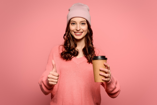 cheerful girl holding coffee to go and showing thumb up on pink background - Foto, afbeelding
