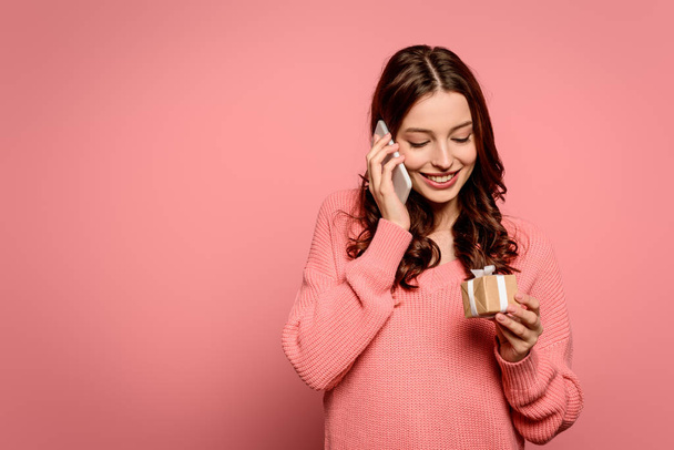 happy girl talking on smartphone and looking at gift box on pink background - Foto, Imagem