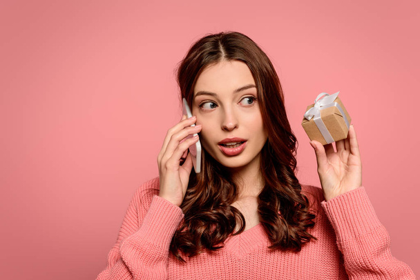surprised girl talking on smartphone and looking at gift box isolated on pink - Photo, Image