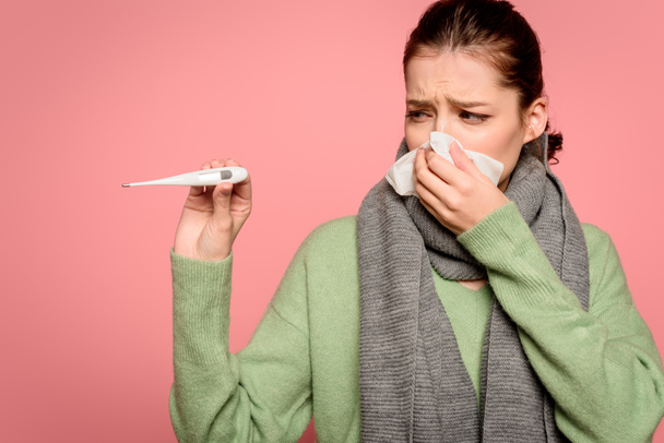 sick girl in warm scarf wiping nose with paper napkin and showing thermometer isolated on pink - Photo, Image