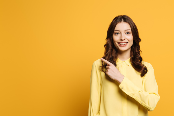 smiling girl pointing with finger while looking at camera on yellow background - Zdjęcie, obraz