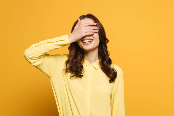 smiling girl covering eyes with hand on yellow background - Foto, afbeelding