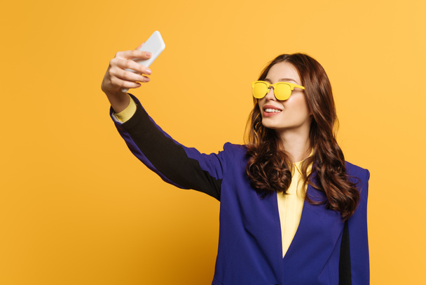 smiling, stylish girl in yellow glasses taking selfie on smartphone on yellow background - Foto, afbeelding