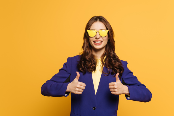 smiling girl in yellow glasses showing thumbs up isolated on yellow - Foto, Imagen