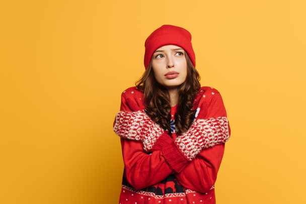 displeased girl in red ornamental sweater and mittens hugging herself while feeling cold isolated on yellow - Φωτογραφία, εικόνα