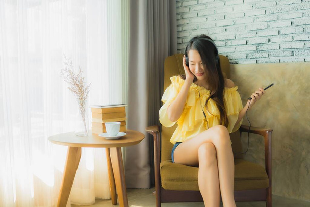 Ritratto giovane donna asiatica seduta sulla sedia ascoltare musica con telefono cellulare caffè e libro in salotto interno
 - Foto, immagini